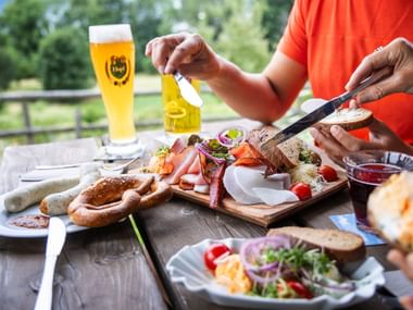 Person in orange shirt preparing traditional Bavarian snack platter with cold cuts, cheese, bread, and beer on wooden table outdoors.
