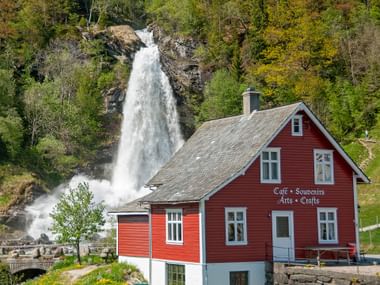 Rotes Holzcafé mit weißen Fenstern vor dem Steinsdalsfossen-Wasserfall bei Norheimsund, umgeben von grünem Wald.
