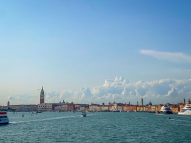 Panoramablick auf die Uferpromenade von Venedig mit Markusturm, historischen Gebäuden und Booten auf türkisfarbenem Wasser unter bewölktem Himmel.