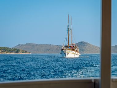 Weißes Segelschiff MS Macek mit zwei Masten auf blauem Wasser, mit felsigen Küsteninseln und Hügeln im Hintergrund unter klarem Himmel.