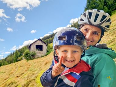 Lächelnde Frau und Kind mit Fahrradhelmen machen ein Selfie auf einem grasbewachsenen Hang mit Holzscheune und Wald im Hintergrund.