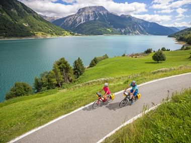 Zwei Radfahrer fahren entlang einer asphaltierten Straße am Reschensee mit türkisfarbenem Wasser, grünen Wiesen und Bergen unter blauem Himmel.