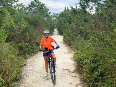 Woman in orange shirt and white helmet cycling on a gravel path through green vegetation in Tuscany under a cloudy sky.