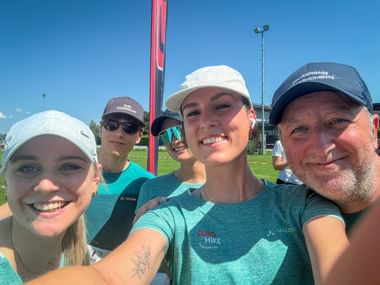 Five people in sports caps taking a selfie at a sports field. They wear turquoise Vaude shirts and smile at the camera under a clear blue sky.