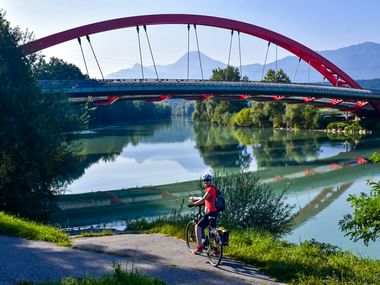 Radfahrer in roter Jacke am Alpe-Adria-Radweg bei moderner roter Bogenbrücke über ruhigem Fluss mit Bergkulisse nahe Villach.