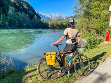 Radfahrer mit Helm steht neben Fahrrad mit gelben Taschen auf Weg entlang der Salzach in St. Johann, Berge im Hintergrund.
