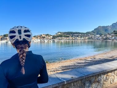 Radfahrerin mit weißem Helm und geflochtenem Haar blickt auf die malerische Bucht von Port de Sóller auf Mallorca mit Booten und Hanghäusern.