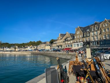 Malerstaffeleien entlang der Uferpromenade von Cancale mit historischen Steingebäuden und blauem Hafenwasser unter klarem Himmel.