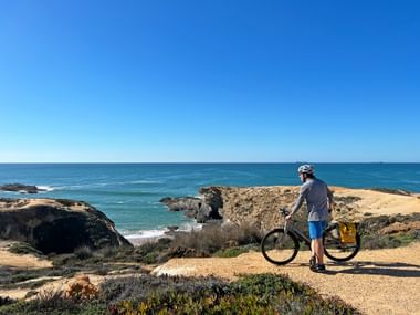 Radfahrer mit Helm auf sandigem Klippenpfad mit Blick auf den Atlantik an der Algarve. Felsformationen und blaues Meer sind zu sehen.