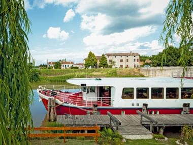 Red and white river cruise ship MS Ave Maria moored at a wooden dock. Willow trees frame the scene with Italian countryside buildings in background.