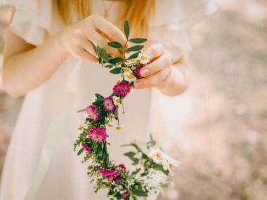 Person in weißem Kleid hält einen handgemachten Blumenkranz mit rosa Blumen, weißen Gänseblümchen und grünem Laub.