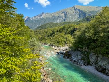 Turquoise Soča River flowing through rocky gorge near Kobarid, framed by green trees with mountain range under blue sky.