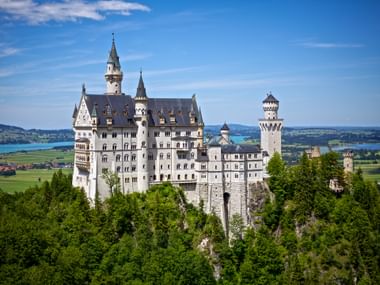 Schloss Neuschwanstein auf einem bewaldeten Hügel mit weißen Türmen. Blaue Seen und grüne bayerische Landschaft im Hintergrund sichtbar.
