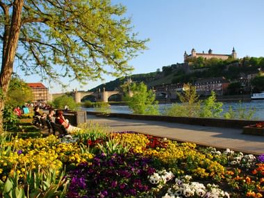 Bunte Blumenbeete entlang des Mains in Bayern mit Menschen auf der Promenade. Festung Marienberg und Steinbrücke im Hintergrund sichtbar.