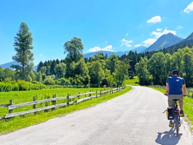 Radfahrer auf asphaltiertem Weg entlang des Drau-Radwegs in Kärnten, mit Holzzaun, grünen Wiesen, Wäldern und Bergen unter blauem Himmel.