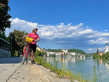 Cyclist in pink jersey with yellow bag standing on riverside path in Passau, with Danube River and historic town buildings under blue sky.