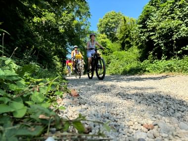Cyclists riding on a gravel path through lush green vegetation near Pradamano on the Alpe Adria cycle route under blue sky.