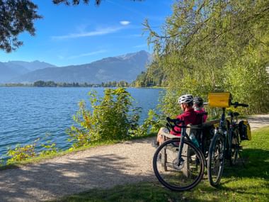 Cyclist in pink jacket resting by Zell am See lake with touring bikes. Mountain panorama and blue sky in background, lush greenery along shore.