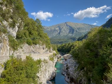Turquoise river flowing through rocky gorge with stone bridge, surrounded by forested cliffs and mountains near Kobarid under blue sky.