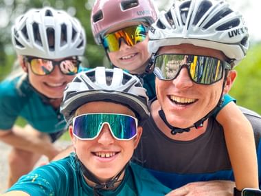 Four smiling cyclists in helmets and sunglasses taking a selfie outdoors. Green landscape visible in background.
