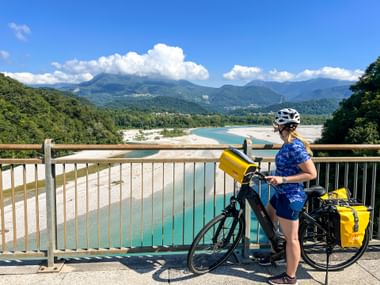 Radfahrerin mit Helm und blauem Trikot steht mit Tourenrad auf der Tagliamento-Brücke in Friaul und blickt auf türkisfarbenen Fluss und Berge.
