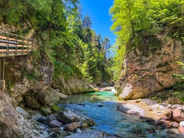 Klarer türkisfarbener Bach fließt durch die felsige Slizza-Schlucht bei Tarvis, mit steilen Felswänden, grünen Bäumen und einem Holzsteg links.