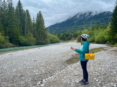 Cyclist with helmet and yellow bag standing on rocky riverbank of Isar River near Mittenwald, with forested mountains and clouds in background.
