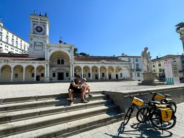 Two cyclists rest on steps in Udine's Piazza Libertà with touring bikes nearby. Historic clock tower and arched colonnade visible under blue sky.
