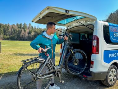 Mitarbeiter in türkisfarbener Jacke lädt Fahrrad in weißen Eurobike-Transporter mit offener Heckklappe. Grüne Wiese und Wald im Hintergrund unter blauem Himmel.