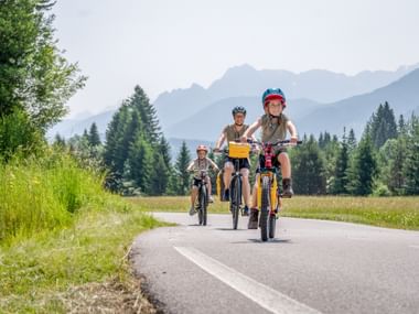 Three cyclists riding on paved path through alpine landscape with mountains, forests and meadows. Child leads on bike with helmet.