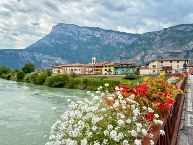 White and red flowers in planters along the Etsch River with San Michele village and mountains in the background.