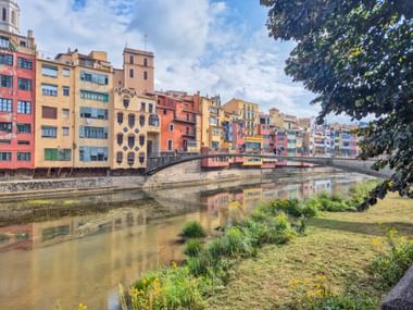 Bunte historische Gebäude in Orange, Gelb und Rot säumen den Fluss Onyar in Girona, Katalonien. Eine Brücke überquert den Fluss mit Vegetation an den Ufern.