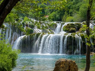 Cascading waterfalls in Krka National Park, Croatia, with moss-covered rocks and turquoise pools framed by lush green trees.