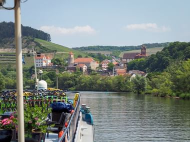 River cruise ship with bicycles on deck moored along the Main River, with the village of Triefenstein and vineyards visible in the background.