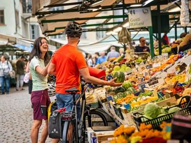 Zwei Radfahrer mit Rädern an einem italienischen Marktstand mit buntem frischem Obst und Gemüse. Schild zeigt 'Gusti Italiani'.