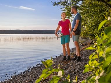 Paar mit Fahrradhelmen am Kiesufer des Starnberger Sees in Bayern, umgeben von grünem Laub mit ruhigem See und Hügeln im Hintergrund.