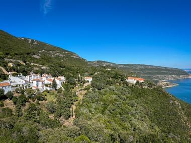 Weiße Gebäude des Klosters Portinho da Arrábida an einem grünen Hang mit Blick auf blaue Küstengewässer unter klarem Himmel.