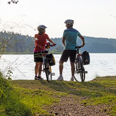 Two cyclists with helmets and panniers standing by their bikes at the shore of Ostersee, viewing the calm lake with forested hills in the background.