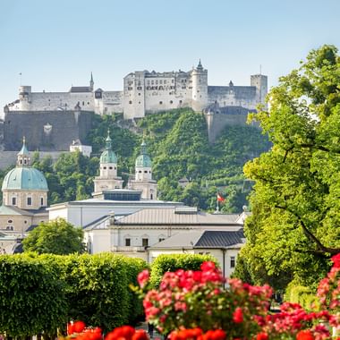 Hohensalzburg Fortress on hilltop overlooking Salzburg's old town with green domes, historic buildings, and red flowers in foreground.