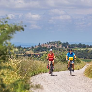 Two cyclists riding on a gravel path through golden fields in Val d'Orcia, Tuscany. A hilltop village with cypress trees is visible in the distance.