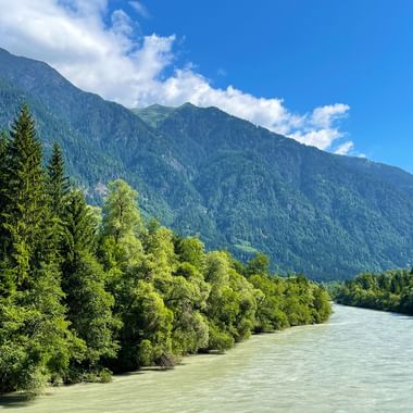 The turquoise Drau River flows through lush green forests with a forested mountain rising under blue sky with white clouds in Austria.