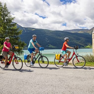 Drei Radfahrer mit gelben Packtaschen fahren am berühmten Kirchturm im Reschensee in Südtirol vorbei, mit türkisfarbenem See und Bergen dahinter.