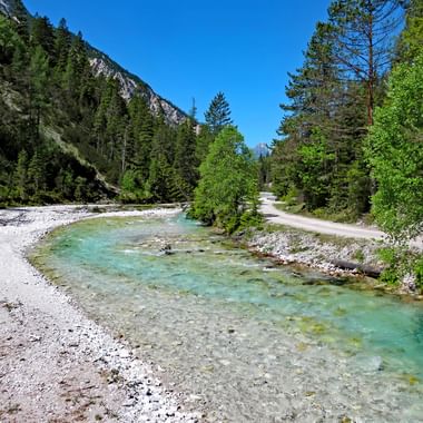 Clear turquoise Isar River flowing through mountain valley with gravel banks, cycling path along the shore, surrounded by green forest and mountains.