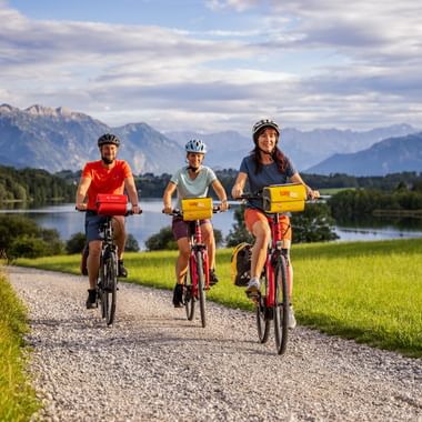 Three cyclists riding on a gravel path near Riegsee in Bavaria, with mountains, lake, and green meadows in the background under a cloudy sky.
