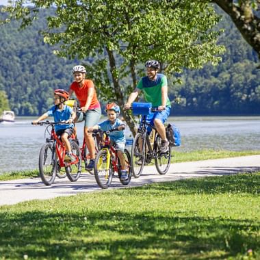 Family of four cycling on path beside the Danube River. Two adults and two children ride bikes under trees with forested hills in background.