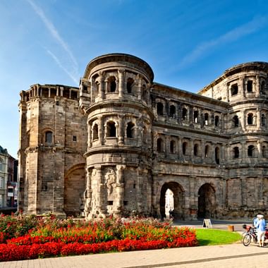 The Porta Nigra Roman gate in Trier with red flower beds in foreground. Cyclists pass by the ancient sandstone structure under blue sky.