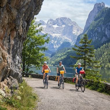 Three cyclists on lakeside path at Gosausee with Dachstein mountain range in background. Rocky cliff on left, alpine lake and forested slopes visible.