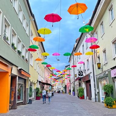 Pedestrian street in Villach old town with colorful umbrellas suspended overhead. Buildings line both sides with shops at street level.