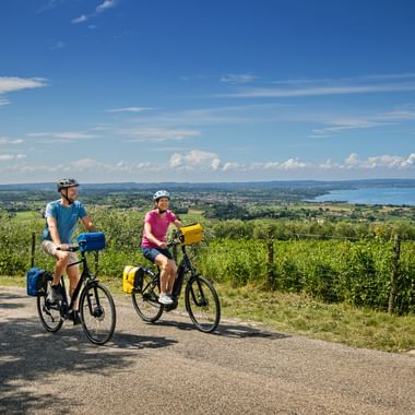 Two cyclists with yellow panniers on a road near Cavaion Veronese, overlooking Lake Garda with vineyards and mountains in the background.