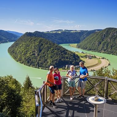 Four people on a viewing platform overlooking the Schlögener Schlinge, a dramatic horseshoe bend of the Danube River surrounded by green hills.